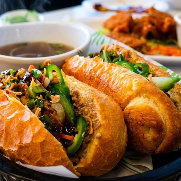 Two banh mi sandwiches with a side of steaming pho soup on a white table.