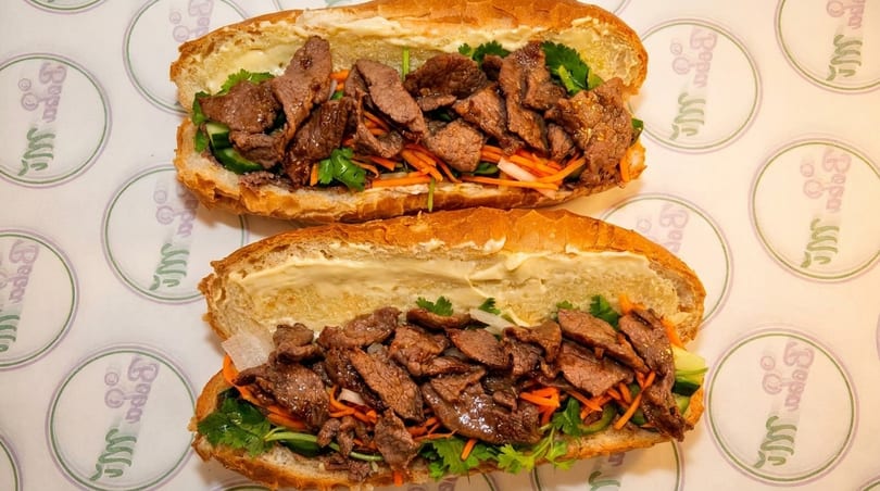Two delicious banh mi sandwiches filled with grilled beef, pickled vegetables, and cilantro on branded paper.