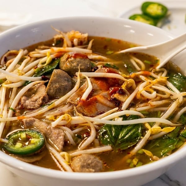 A white bowl of pho with meat, bean sprouts, jalapeños, and a spoon on a marble table.
