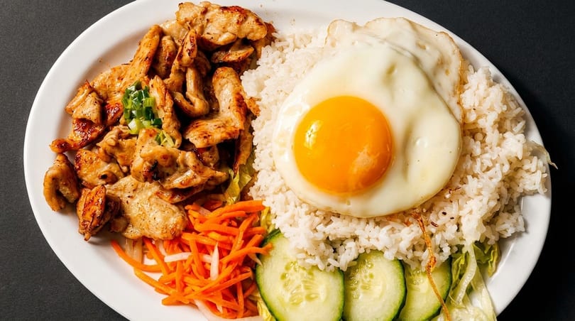 Grilled chicken, white rice, fried egg, pickled carrots, and cucumber slices on a white plate.